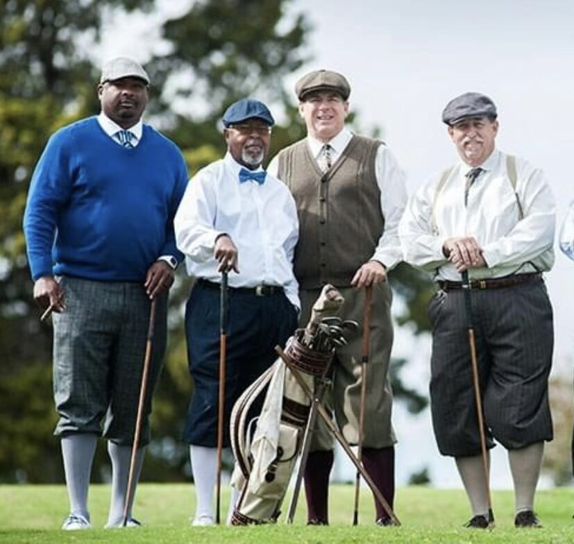 Four golfers in knickers, long socks, and caps standing around a vintage golf bag of hickory clubs